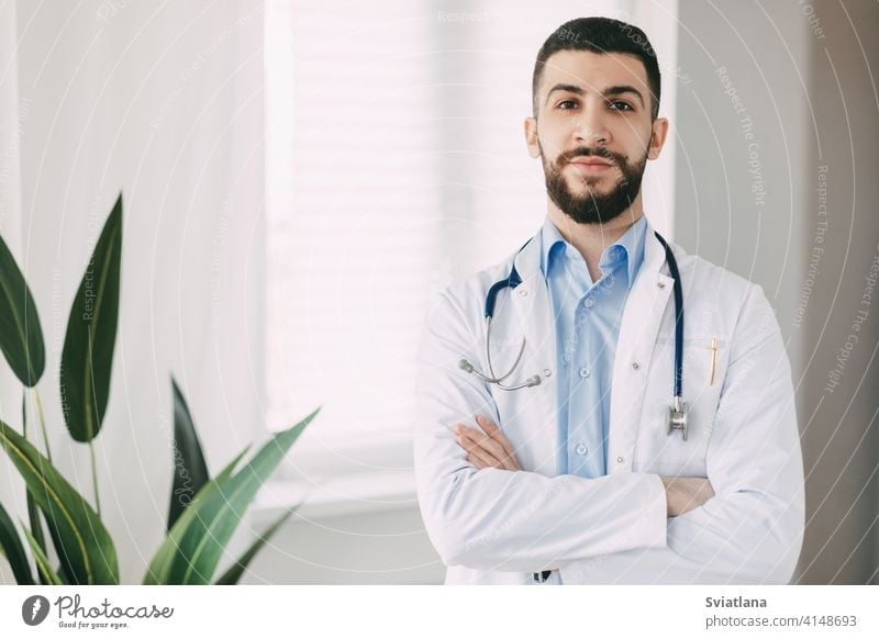 Smiling young doctor with stethoscopes and white medical gown stands in his office with arms crossed medicine man health professional cheerful male healthcare