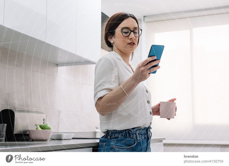 woman drinking coffee in the kitchen while using her smartphone indoor one person relaxation young adult apartment comfortable routine relaxing cozy brunette