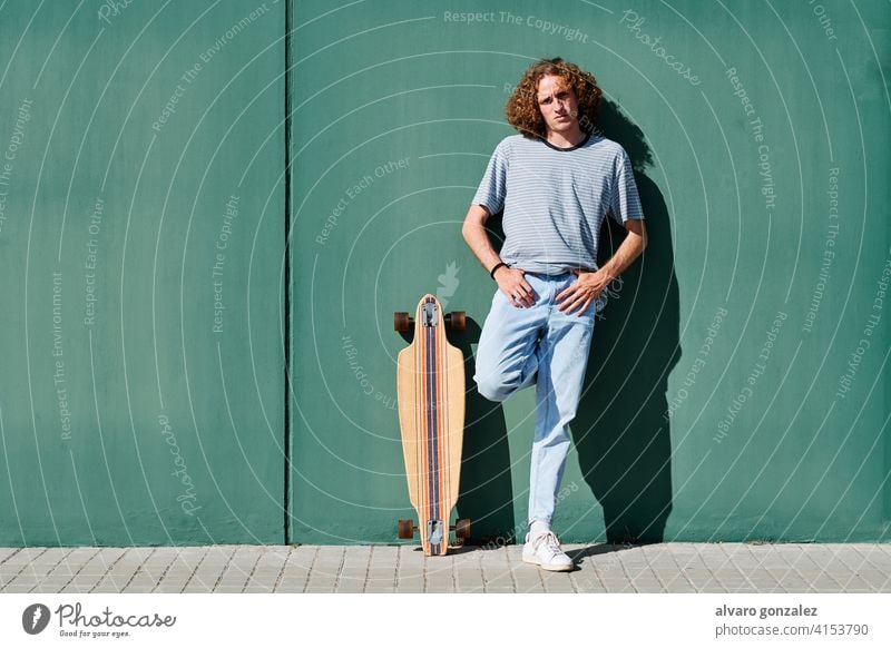 a young man with curly hair standing on a green wall on a sunny day with his skate or longboard che skateboard attractive person skateboarding sport skater male