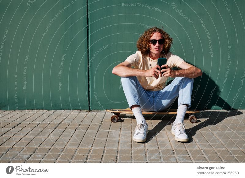 a young man using his smartphone and seated on his skateboard or longboard with a wall behind him che attractive person skateboarding sport skater male guy