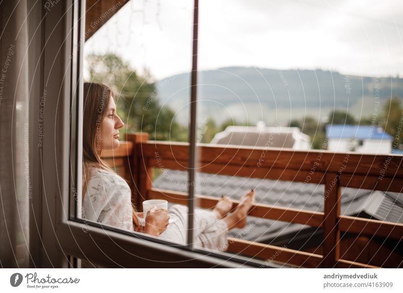 Young female standing after taking a shower in the morning on balcony of the hotel. holding a cup of coffee or tea in her hands. Looking outside nature forest and Mountain