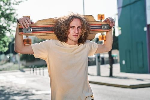 young man with curly hair looking at camera with his longboard or skeateboard on his shoulders in the middle of the street che skateboard attractive person