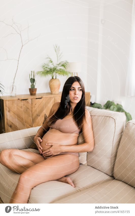 Smiling pregnant woman in sofa at home touch belly pregnancy cozy lingerie underwear gentle tummy female delicate couch mother care tender sit expect parent