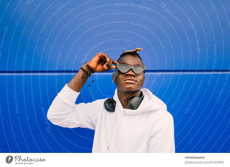 Young afro American black man wearing a white sweatshirt, sunglasses, comb and headphones on a blue wall with confidence young african street background cool