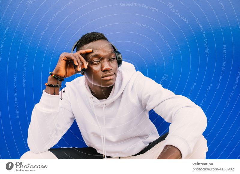 Young afro American black man wearing a white sweatshirt and headphones seated on a blue wall looking to the side. Modern look young african street background