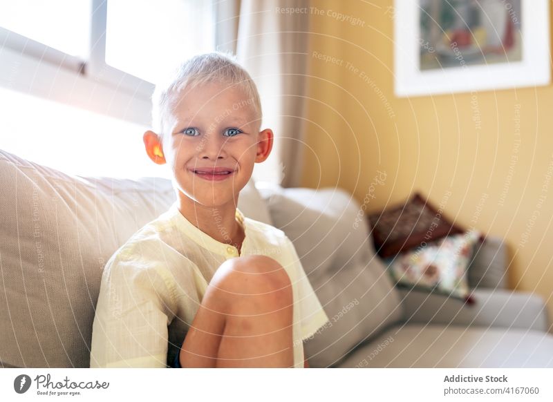 Positive child smiling while resting on sofa at home relax couch smile cheerful free time positive adorable childhood joy comfort kid blue eyes blond delight