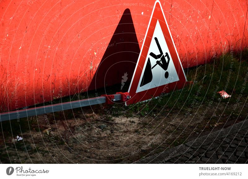 End of work at the construction site: Construction workers have laid the warning sign with a black pictogram framed in red on a white background flat on the sandy ground in front of a red construction fence illuminated by the shadows cast by the evening sun.