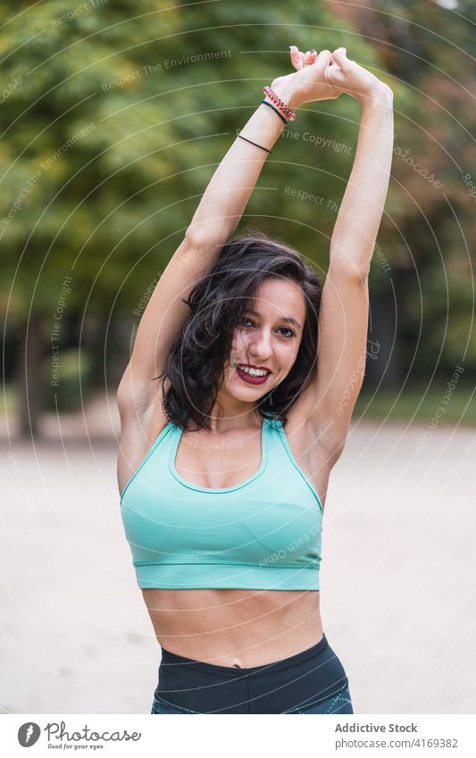 Smiling sportswoman warming up arms during workout in park warm up stretch training flexible athlete fitness female bra healthy exercise above head arms raised