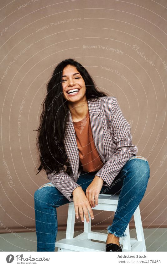 Happy ethnic woman sitting on chair in studio trendy smart casual style happy model jacket smile outfit cheerful confident female laugh fashion contemporary