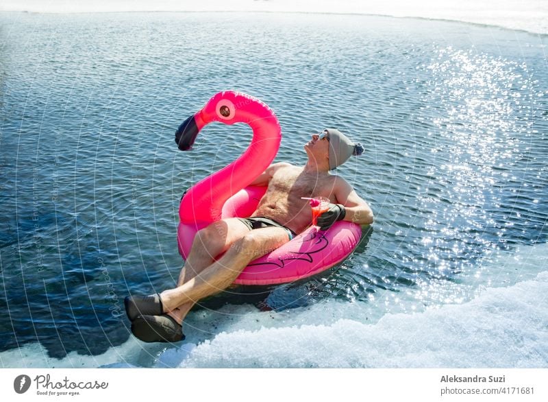A man swimming in an ice hole in winter in Finland, floating on a pink inflatable flamingo with cocktail in hand. Vacation options, dreaming of summer. active