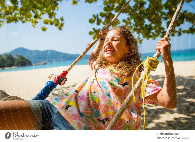 Happy cute little girl laughing and swinging on swing on the tree at the beach. Beautiful summer sunny day, turquoise sea, rocks, white sand, picturesque tropic landscape. Phuket, Thailand. Carefree
