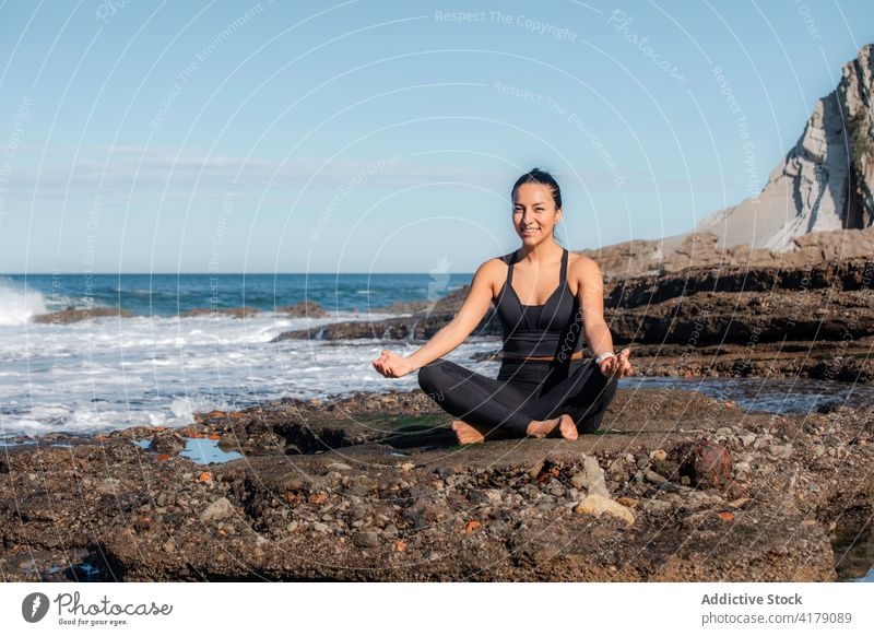 Relaxed woman doing yoga on seashore beach meditate lotus pose asana mudra padmasana female legs crossed zen nature wellbeing harmony coast energy slim cheerful