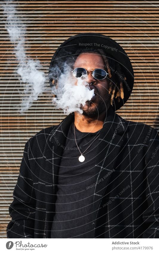 Trendy black man in hat smoking smoke exhale trendy cool lifestyle modern contemporary dreadlocks sunglasses african american ethnic personality street style