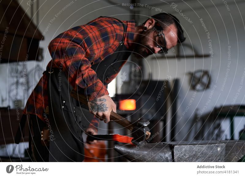 Focused young blacksmith heating and striking metal on anvil in workshop man strike iron smithy spark manual hammer craftsman male beard brutal apron goggles