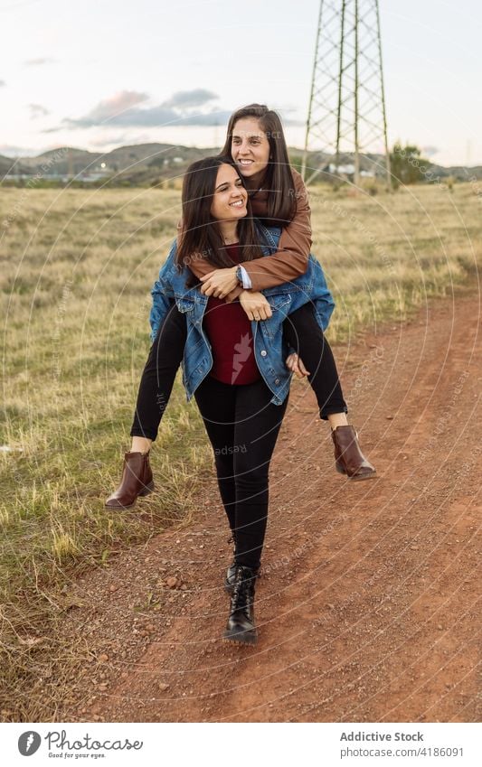 Delighted lesbian couple having fun in nature piggyback women ride cheerful girlfriend lgbt relationship together weekend road smile homosexual love fondness