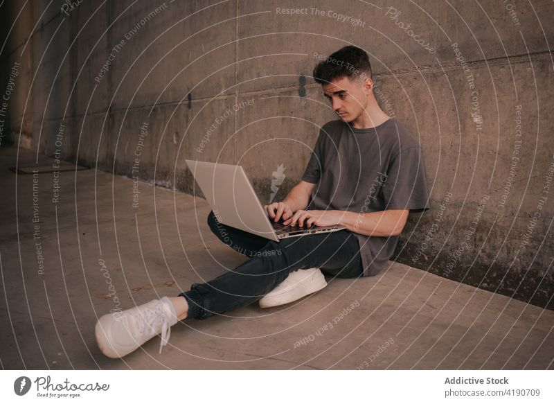 Concentrated young male freelancer working distantly on laptop man internet serious social media concentrate job trendy browsing lean on concrete wall