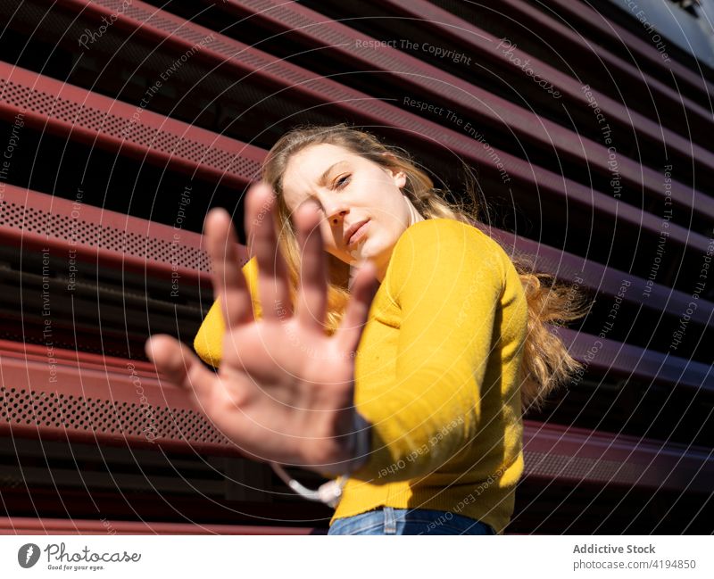 Contemplative young woman reaching out hand towards camera on city street reach out pensive thoughtful calm touch hair appearance style sunlight facade fence