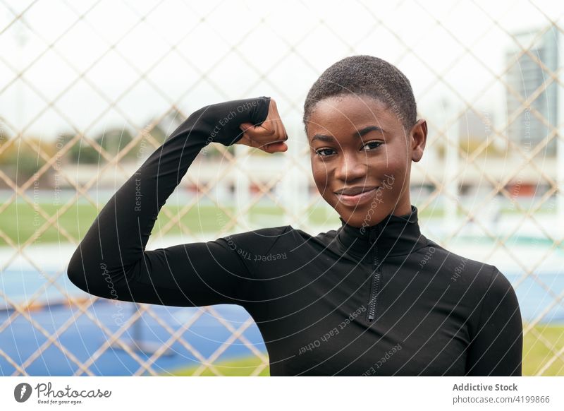 Cheerful black woman showing muscle against grid fence strong power smile feminine gentle candid town portrait gesture sportswoman energy wellness modern style