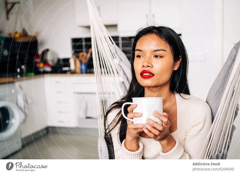 Hispanic lady sitting in hammock at home woman beverage female hispanic young drink tea mug free time cup apartment contemporary daytime indoors wistful