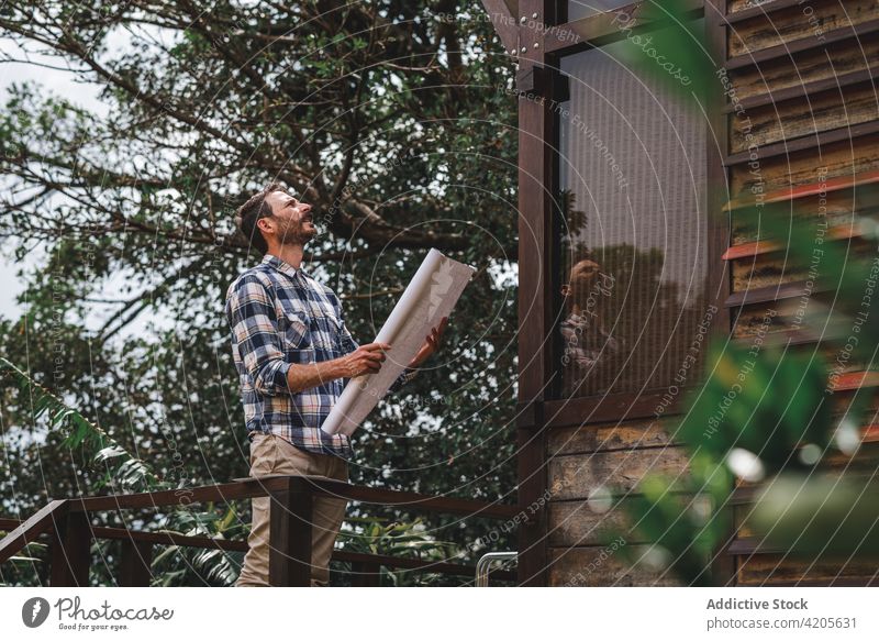 Male architect with blueprint standing near modern house man building project draft paper terrace wooden male cottage construction forest guy estate