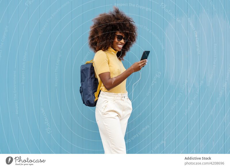 Cheerful black woman browsing smartphone near blue wall selfie moment content headphones portrait using gadget colorful device self portrait happy spare time