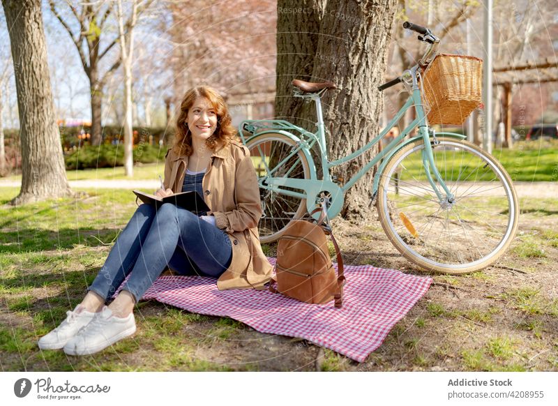 Happy young woman writing in planner on lawn near bicycle happy write notebook park take note checkered fabric female thoughtful hobby casual concentrate calm