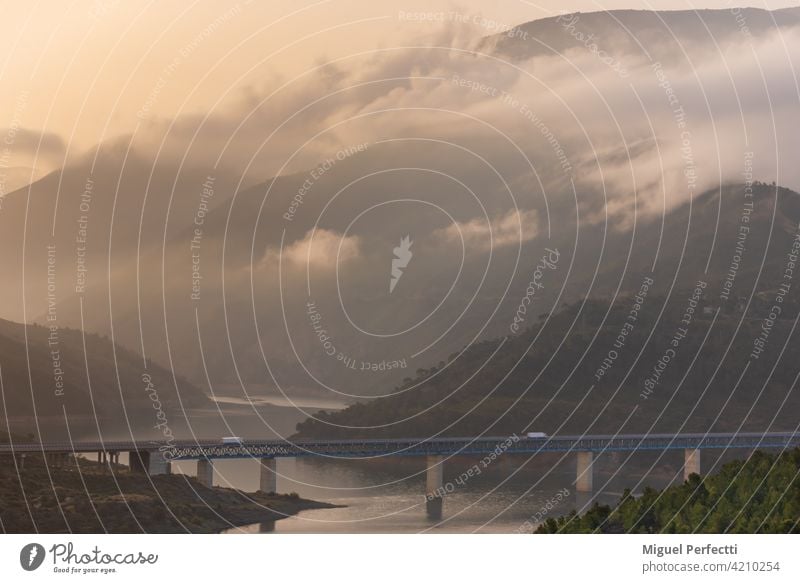 Refrigerated trucks driving on a bridge over a swamp surrounded by mountains and forests with low clouds at sunrise. landscape nature water lake river