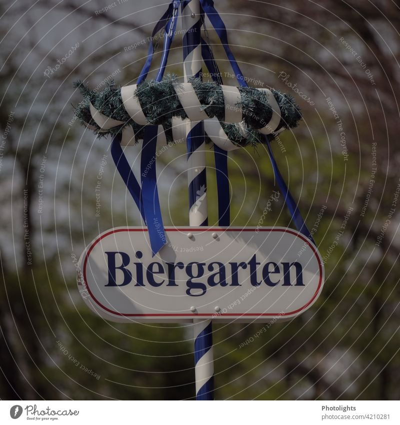 Sign "Biergarten" is attached to a maypole with a wreath and blue ribbons. May tree Beer garden sign Wreath tapes Blue Green White Deserted Colour photo