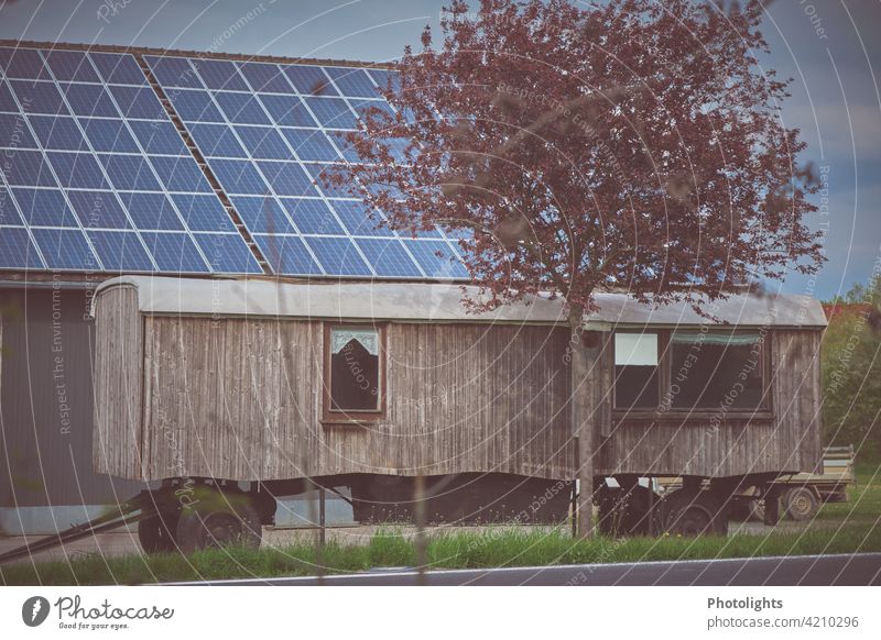 Wooden caravan stands in front of a barn with solar roof. Flowering tree in the foreground. Site trailer Trailer Caravan Barn Drape Wheels trailer coupling Tree