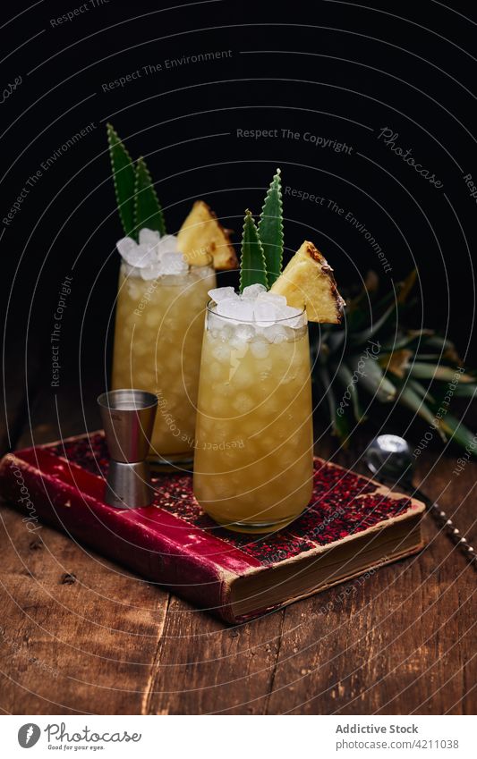 Glasses with cocktails with pineapple pieces on book glass ice refreshment table leaf drink spoon beverage fruit alcohol tasty cold yummy portion ingredient