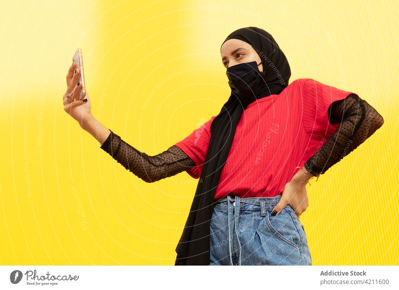 Trendy Muslim woman in face mask taking selfie on smartphone self portrait trendy fashion cool individuality hand on hip using gadget device modern vivid wear