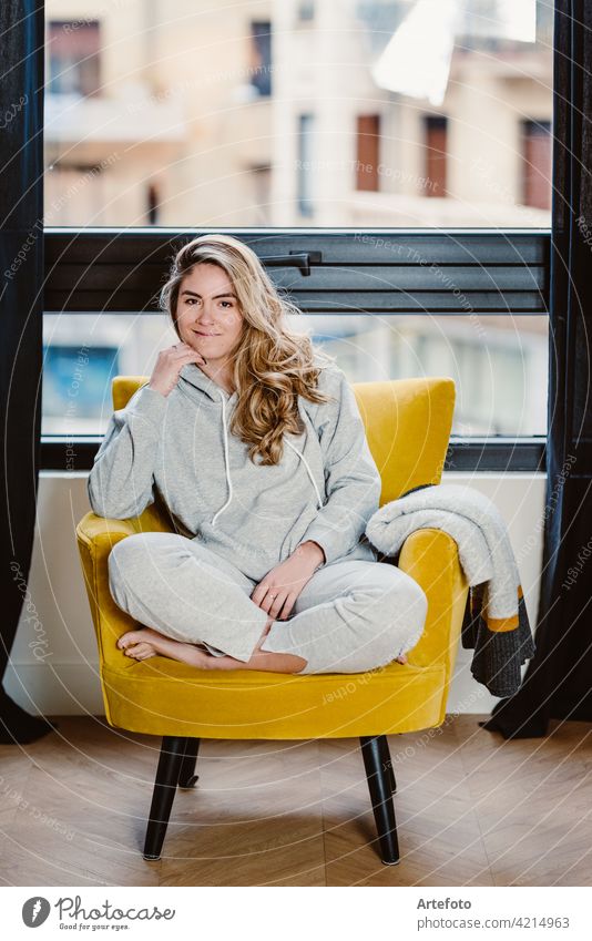 Portrait of a beautiful blonde girl sitting in a yellow sofa and wearing sportswear looking at camera. portrait woman smile couch young down happy home room