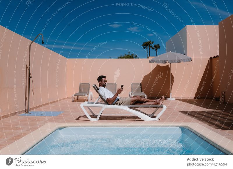 Man lying on deckchair and browsing smartphone near pool man freelance poolside telework summer lounger male surfing mobile internet online using remote gadget