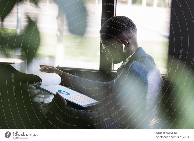 Serious black businessman in earbud with diagram on paper indoors work project startup serious clipboard cafeteria using gadget device executive tws