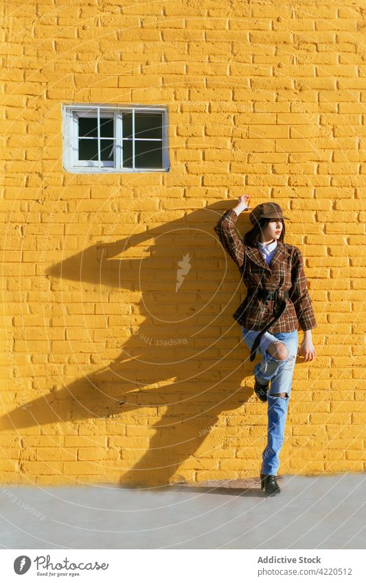Trendy model in ripped jeans leaning on painted brick wall style fashion individuality leg raised shadow woman street pavement stylish window yellow color