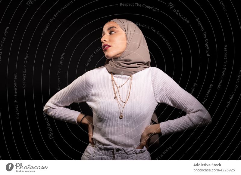 Stylish Muslim woman standing in dark studio muslim headscarf style modish confident appearance outfit headdress garment posture culture model young feminine