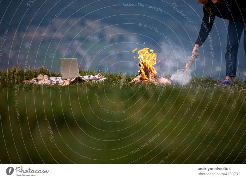 young woman freelancer throws firewood into a fire in the mountains in the evening. tourist girl resting and working outdoor female laptop sitting outside