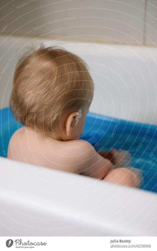 11 month old baby with back to camera sitting in a blue plastic bath tub; blonde wispy hair bath time clean reach grasp water play back of head baby hair seated