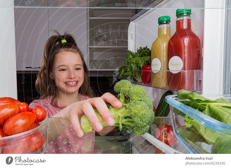 Open fridge from the inside, glass shelves with colourful vegetables, bottles of organic juices. Cute little girl with happy smile taking broccoli. Healthy eating, vegan concept.