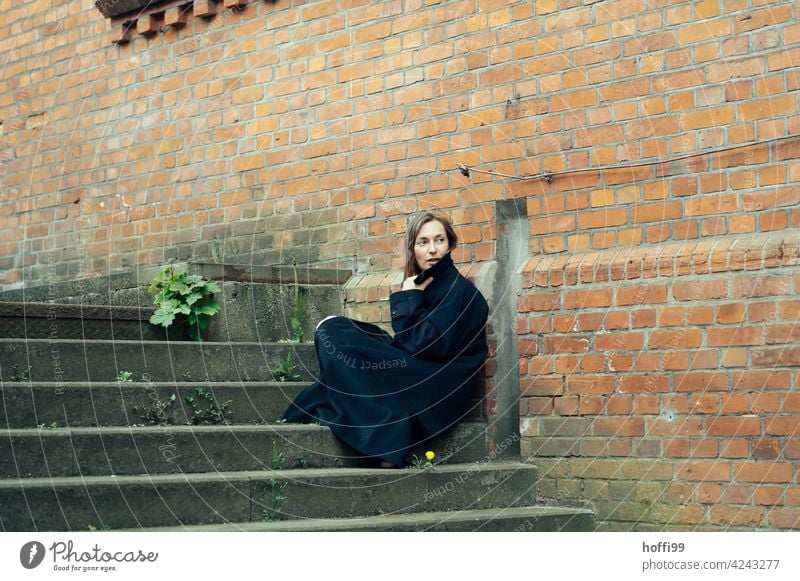 the woman sits on a staircase wrapped in a cloak and looks back the way over her shoulder Young woman Coat melancholy Face of a woman Feminine long hair