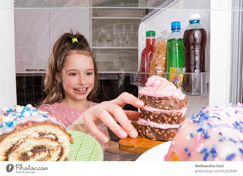 Open fridge from the inside, glass shelves with colourful desserts, cakes, cookies, candies, bottles of sugar drinks. Unhealthy eating, sugar food concept. Little girl with happy smile taking cake