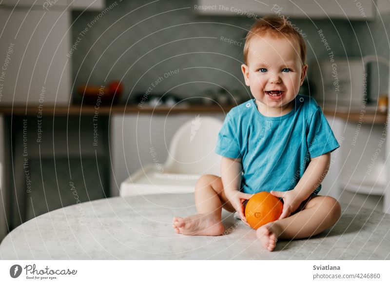 A little boy sits on the kitchen table with an orange in his hands and smiles baby eating kid healthy happy fresh toddler cute food child citrus fruit nutrition