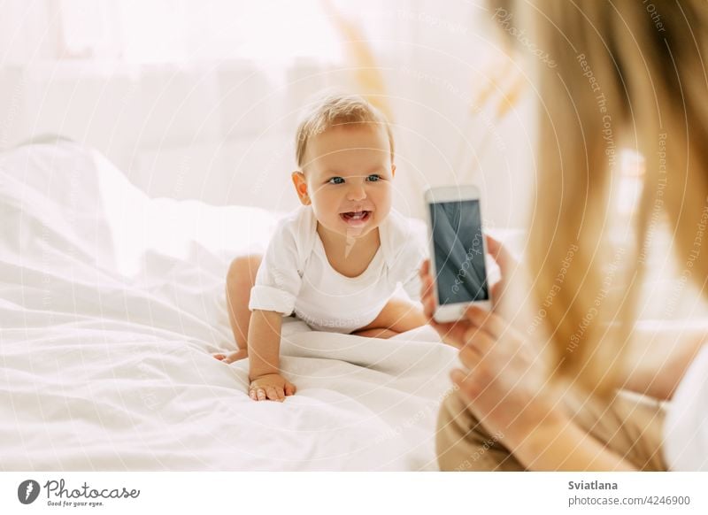 Cute baby is sitting on the bed and posing for his mom, mom is taking pictures of her baby on her mobile phone. Modern technologies, space for text beautiful
