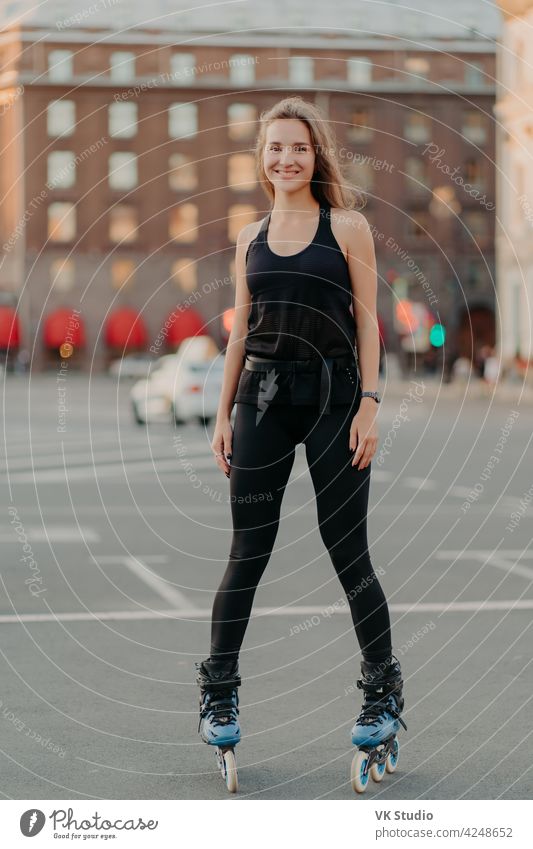 Positive young female model in sportsclothes rides on blades enjoys leisure activities poses at urban place against blurred background stands in full length. Active lifestyle and rollerblading concept