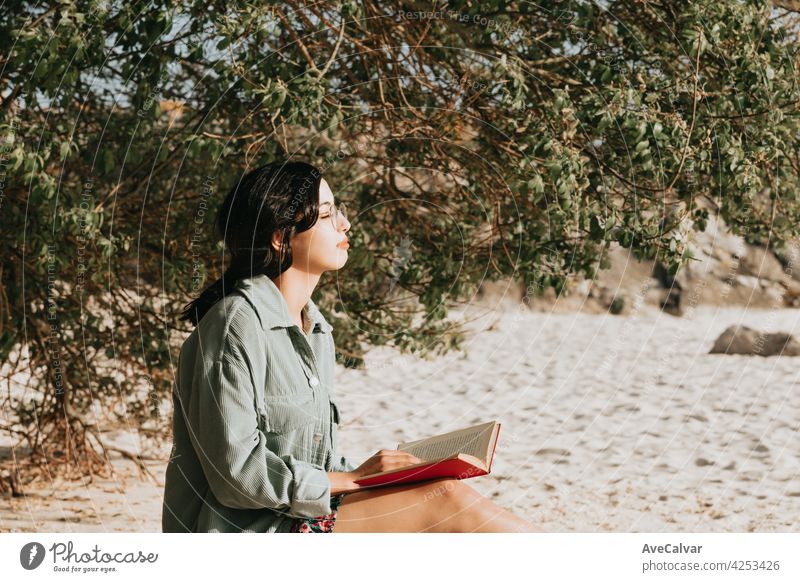 Young Moroccan woman on modern clothes sitting on the beach reading a book during a sunny day with copy space inspirational and relax theme with colorful tones