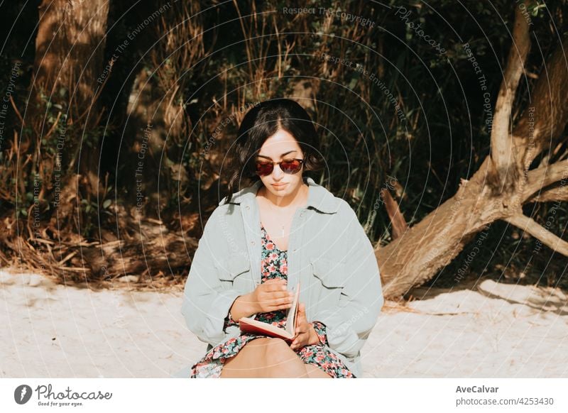 Young Moroccan woman on modern clothes using sunglasses sitting on the beach reading a book during a sunny day with copy space inspirational and relax theme with colorful tones