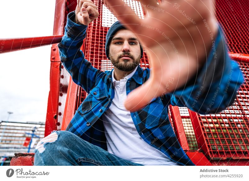 Man in blue hat and checkered shirt near metal construction man iron confident fist show gesture cool male beard style casual appearance personality outfit guy