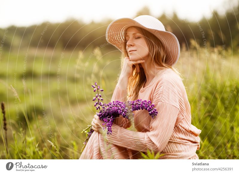 Portrait of a beautiful blonde with a bouquet of lupines in her hands sitting on the lawn girl field hat summer young dress flower beauty portrait