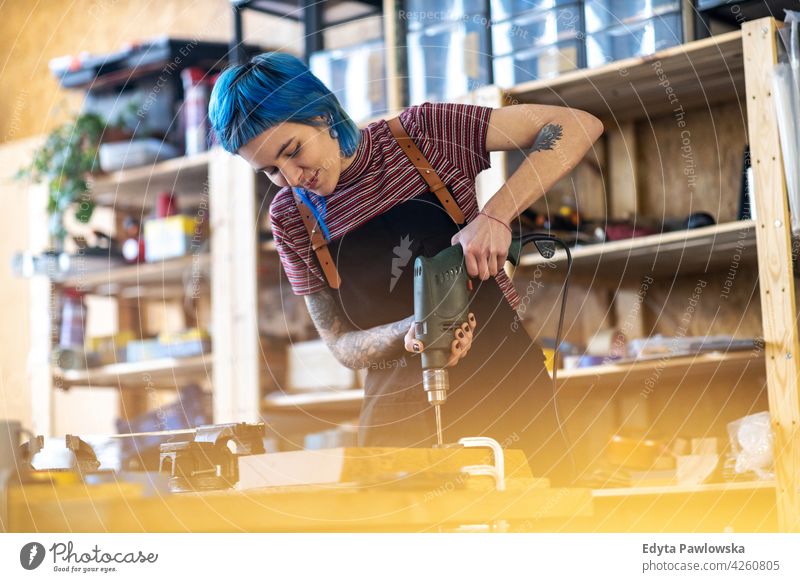 Young woman using electric drill in industrial workshop diy hipster colorful hair tattoos female owner profession service small business employee working