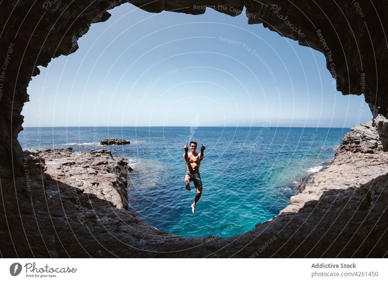 Smiling man showing fuck gesture while jumping over sea ocean cave content nature highland hole sky activity energy smile rude middle finger rebel aggressive